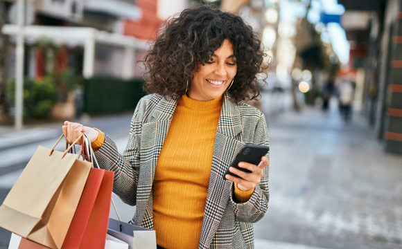 A woman on the street with some new purchases in one hand and a phone in the other.