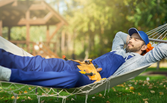 A man in overalls and a toolbelt resting in a hammock on a fall day.